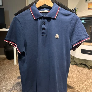 Mörkblå pikétröja från Moncler - Säljer en mörkblå pikétröja från Moncler med klassisk krage och knappar framtill. Tröjan har röda och vita ränder på krage och ärmslut samt Moncler-logga på bröstet. 