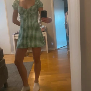 Grön och vit rutig kortklänning med puffärmar - Superfin kort klänning i grönt och vitt rutigt mönster. Klänningen har puffiga korta ärmar, söt urringning och knappar framtill. Perfekt för sommarens alla tillfällen. Materialet känns lätt och luftigt.