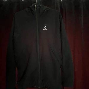 Svart vindjacka från Haglöfs - Säljer en svart vindjacka från Haglöfs med dragkedja framtill och diskret logga på bröstet. Jackan har huva och två sidofickor, perfekt för blåsiga dagar. Enkel och stilren design som funkar till det mesta. Storleken är M men passar även personer som använder L 