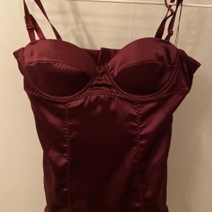 Vinröd korsettopp från Victoria's Secret - Säljer en vinröd korsettopp från Victoria's Secret i glansigt satinmaterial. Toppen har vadderade kupor, justerbara axelband och en söt volangkant nertill💕💕