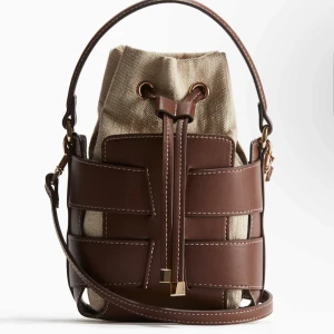Brun bucket bag med canvasdetaljer - Snygg brun bucket bag med detaljer i canvas och justerbar axelrem. Väskan har ett unikt flätat mönster i brunt skinn och stängs med snörning. Perfekt accessoar för att lyfta din outfit.