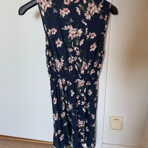 Mörkblå blommig klänning  - Säljer en mörkblå klänning med rosa och vita blommor. Klänningen är ärmlös, med resår i midjan för en bekväm passform. Perfekt för varma dagar och somriga tillfällen.