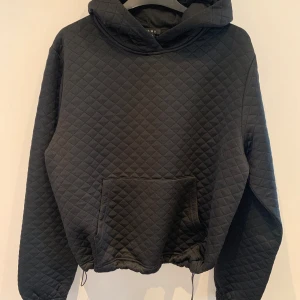 Svart quiltad hoodie från Amisu - Säljer en svart quiltad hoodie från Amisu med huva och magficka. Tröjan har långa ärmar och dragsko nertill för justerbar passform. Perfekt för en avslappnad och trendig stil.
