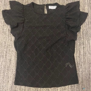 Svart blus med volangärmar - Säljer en svart blus från Holly & Whyte by Lindex. Blusen har korta volangärmar och ett fint mönster. Man ser igenom hålen i mönstret på blusen men det är bara att ha något linne under. Bara använd två gånger🫶🏼