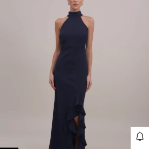 Mörkblå långklänning med volang - Elegant mörkblå långklänning med halterneck och volangdetalj framtill. Klänningen har en hög halsringning, är ärmlös och har en slits framtill med volang. Perfekt för festliga tillfällen.