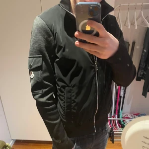 Moncler - Säljer en svart jacka från Moncler med klassisk patch på ärmen och dragkedja framtill. Jackan har ficka med knapp på ärmen och ribbade muddar. Insidan har ett unikt serietidningsmönster. Perfekt för dig som vill ha en stilren och exklusiv look.