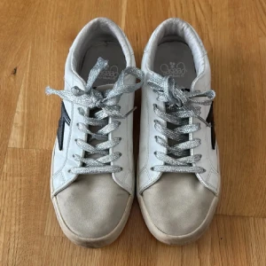 Unika skor  - Säljer ett par vita sneakers från Le temps des cerises  med silverfärgade skosnören och svart blixtdetalj på sidan. 