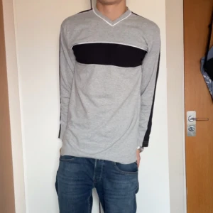 Grå långärmad tröja med svart detalj - En grå långärmad tröja i s med svart panel över bröstet och svarta detaljer längs ärmarna. Tröjan har v-ringad hals med unik design. Killen är 184 cm och väger 73 kg