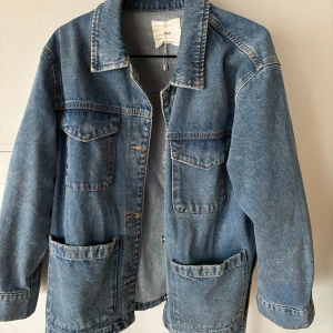 Blå jeansjacka med bälte - Snygg blå jeansjacka med knappar och praktiska fickor fram. Jackan har ett bälte i midjan för en justerbar passform och klassisk krage. Perfekt för våren. Är i storlek L men passar mig som är en S/M
