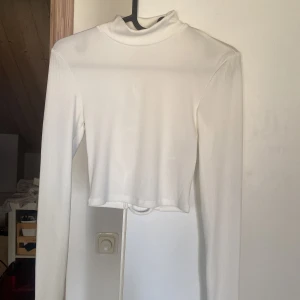 Vit långärmad croppad topp från Amisu - Säljer en vit långärmad croppad topp från Amisu med hög hals och snygg snörning i öppen rygg. Toppens material är ribbad och stretchigt,