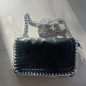 Svart axelväska med silverkedja - Cool svart axelväska i skinn med grov silverkedja som axelrem och detaljer runt kanterna. Perfekt för att lyfta vilken outfit som helst med en edgy touch. Väskan har en rektangulär form och känns både trendig och unik. (Inte min bild❤️)Minsta storleken från tiamo