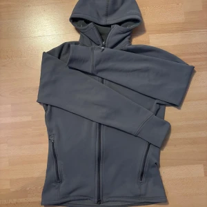 Houdini Hoodie - Mörkgrå fleecetröja från Houdini med huva och hel dragkedja framtill. Tröjan har två sidofickor med dragkedja och är långärmad. Perfekt för lager-på-lager och utomhusaktiviteter. Ny skick 10/10 strl M. 