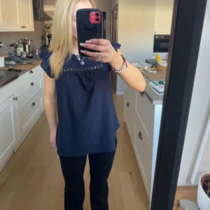 Mörkblå blus med volangärmar - Säljer en mörkblå blus med korta volangärmar och broderade detaljer upptill. Blusen har en lös passform och är perfekt för dig som gillar en söt och stilren look.