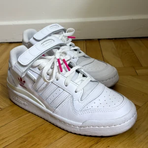 Vita Adidas sneakers  - Säljer ett par fräscha vita Adidas sneakers med klassiska tre ränder, kardborreband och snörning. Skorna har rosa detaljer på snörningen och Adidas-logga på sidan och innersulan. Perfekta för dig som gillar en clean och sportig stil. De är använda 3-4 gånger. 