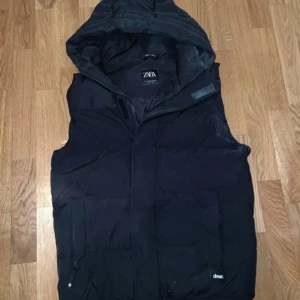 Svart dunväst med huva från Zara - Säljer en svart dunväst från Zara med huva och dragkedja framtill. Västen har en enkel och stilren design, perfekt för lager-på-lager. Passar bra till kyligare dagar och har praktiska fickor.