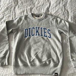 Grå sweatshirt från Dickies - Säljer en klassisk grå sweatshirt från Dickies med blå och vit logga tryckt över bröstet. Tröjan har rund halsringning, ribbade muddar och är tillverkad i mjuk bomullsblandning. Perfekt för en avslappnad stil. Ganska stor is storleken men passar mer som S/M