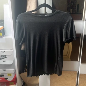Svart ribbad t-shirt från 157 - Svart ribbad t-shirt från 157 med tight passform. T-shirten har korta ärmar och vågiga kanter vid ärmslut och nederkant för en extra detalj. Perfekt för en stilren och enkel look.
