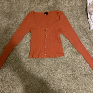 Rostfärgad ribbad topp med knappar från Gina Tricot - Säljer en snygg rostfärgad långärmad topp från Gina Tricot. Toppen är ribbad, croppad och har dekorativa knappar framtill. Perfekt till jeans eller kjol för en trendig look.