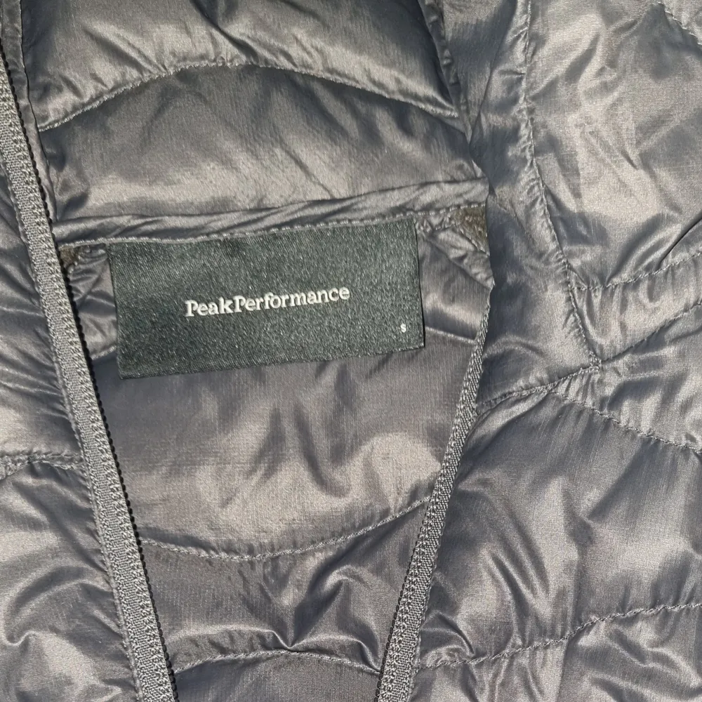 Mörkgrå jacka från Peak Performance med quiltad design och dragkedja. Jackan har långa ärmar och en smidig huva.. Takit.