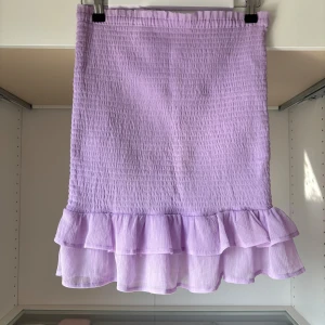 Ljuslila volangkjol från Bik Bok - Supersöt ljuslila kjol från Bik Bok med smockad midja och dubbla volanger nertill. Kjolen är kort och har en luftig känsla, perfekt för varma dagar. Enkel att matcha med olika toppar för en trendig look.