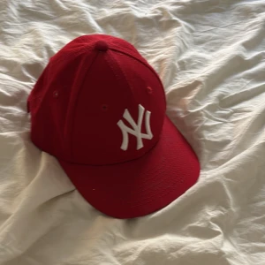 Röd New Era keps med NY-logga - Snygg röd keps från New Era med vit NY-logga framtill. Klassisk modell med böjd skärm och justerbar passform baktill. Perfekt accessoar för en sportig look.