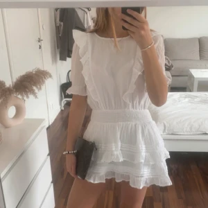 Vit student/skolavslutningsklänning - Supersöt vit volangklänning från Gina Tricot med volangdetaljer och smock i midjan. Klänningen har korta ärmar, rund halsringning och flera lager volanger på kjolen för en luftig känsla. Perfekt för sommarens alla tillfällen!