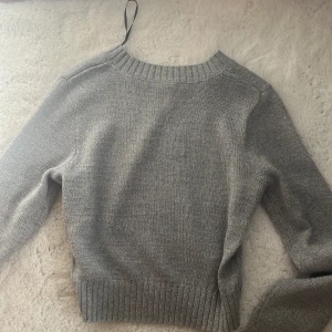 Grå stickad tröja - Säljer en stilren grå stickad tröja med rund hals och långa ärmar. Perfekt för kyligare dagar med ribbade muddar vid ärmslut och nederkant. Enkelt att matcha med olika outfits.