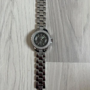 Silverfärgad klocka med stenar - Säljer en snygg silverfärgad klocka från  Michael Kors med länkarmband i metall och glittrande stenar runt urtavlan. Klockan har svart urtavla med tydliga siffror och datumvisning. Perfekt accessoar för att lyfta din stil.