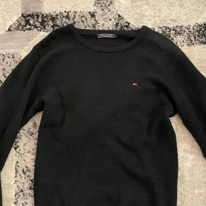 Svart stickad tröja från Tommy Hilfiger - Säljer en svart stickad tröja från Tommy Hilfiger med diskret logga på bröstet. Tröjan har rund halsringning och lång ärm, perfekt för en stilren och enkel look.