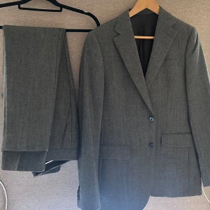 Grå kostym med kavaj och byxor - Elegant grå kostym med en klassisk kavaj och matchande byxor. Kavajen har två knappar och en stilren design, perfekt för formella tillfällen. Byxorna är i samma grå nyans och ger en enhetlig look. Nyskick o endast använd 2 gånger. Nypris: 3000