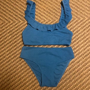 Blå bikini med volangdetaljer - Snygg blå bikini med volangdetaljer på axelbanden. Överdelen har en ribbad textur och ger en trendig look. Perfekt för sommarens stranddagar!longa axel band 