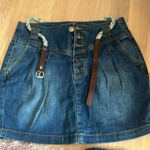 Jeanskjol med bälte - Snygg jeanskjol i blå denim med knappar framtill och ett flätat bälte i midjan. Kjolen har fickor och dekorativa sömmar som ger en cool look. Perfekt för en avslappnad stil.