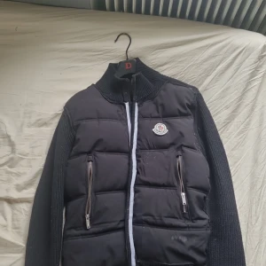 Svart pufferjacka från Moncler - Säljer en svart pufferjacka från Moncler med stickade ärmar och hög krage. Jackan har två framfickor med dragkedja och en broderad Moncler-logga på bröstet. Perfekt för dig som vill ha en snygg och varm jacka med sportig känsla. Jackan är gjord med bomullstråd och fint/dyrt material. Processen har gått långt smidigt bra! Priset är p grund av att jackan är dunder inte för märket!!)