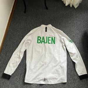 Vit träningsjacka från Craft med Bajen-tryck - Säljer en vit träningsjacka från Craft med grönt Bajen-tryck på ryggen och klubbmärke på bröstet. Jackan har hel dragkedja, svarta muddar och fickor framtill. Perfekt för fotbollsträning eller som supporterplagg.