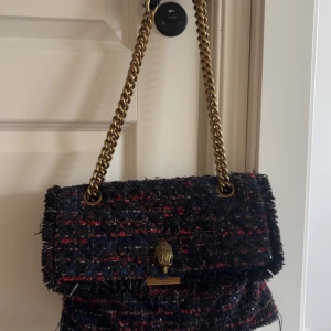 Mörkblå axelväska från Kurt Geiger med kedjerem - Säljer en snygg axelväska från Kurt Geiger i mörkblått tyg med inslag av rött och guld.  Den är i bra skick och man får plats med mycket saker i den!