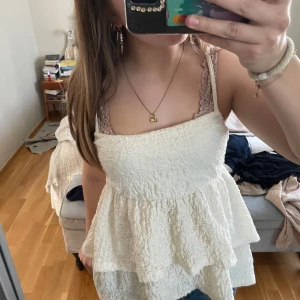 Volang topp gina tricot  - Säljer en söt vit topp från Gina Tricot med volanger och smockdetaljer. Toppen har smala axelband och en luftig passform, perfekt för varma dagar. Den är både stilren och bekväm, vilket gör den till ett mångsidigt tillskott i garderoben.