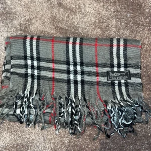 Burberry Halsduk - Säljer en klassisk grå rutig halsduk från Burberry i 100% lammull. Den har ett tidlöst mönster med inslag av svart, grå och rött. Halsduken har fransar i ändarna som ger en extra touch. Perfekt för att hålla dig varm och stilren under kyliga dagar.