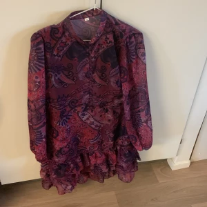 Mönstrad volangtunika i lila blått och rött lote rosa - Säljer en färgstark kortklänning med volangkant och lång ärm. Klänningen har ett paisleyinspirerat mönster i lila, rött, blå och rosa toner. Lätt transparent material och knäppning framtill. Perfekt för dig som gillar unika plagg med mycket färg. Volang nertill! svart underkjol ner till och rekommenderar linne under eftersom att den är lite genomskinlig. Kort klänning men mee som en tunika