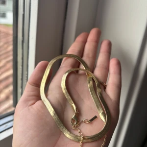 Guldigt halsband från Pilgrim - Säljer ett snyggt guldigt halsband från Pilgrim med platt, ormliknande kedja. Aldrig använt, fortfarande i orginalpaketet. Endast använt för att visa storlek. Första bilden är på minsta storleken, andra bilden är på mellersta storleken. Halsbandet har justerbar längd och stängs med karbinhake. Perfekt för att ge din outfit en trendig touch. Originalpris 399kr