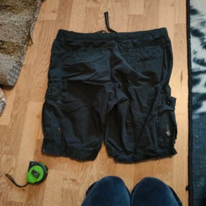 Svarta cargoshorts med snörning - Svarta cargoshorts med flera fickor på sidorna och snörning i midjan. Klassisk cargomodell med avslappnad passform, perfekt för varma dagar. Shortsens material känns slitstarkt och de har en enkel design utan synliga mönster.