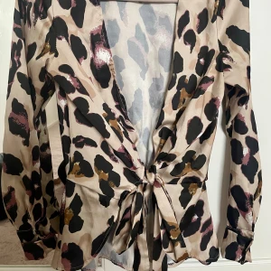 Leopardmönstrad omlottblus med knyt - Säljer en beige omlottblus med svart, brun och rosa leopardmönster. Blusen har långa ärmar, knyt i midjan och lätt glansigt material. Perfekt för dig som gillar djurmönster och vill sticka ut. Storlek S, kan vara liten in storlek. 