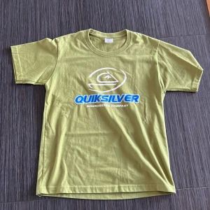Grön t-shirt från Quiksilver - Säljer en grön t-shirt från Quiksilver med tryck i vitt och blått på framsidan. T-shirten har en rund halsringning och korta ärmar. Perfekt för en avslappnad stil. Pris kan diskuteras.