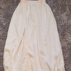 Beige långkjol från Gina Tricot - Säljer en elegant beige långkjol från Gina Tricot. Kjolen har en elastisk midja för en bekväm passform och är tillverkad i ett lätt och flödande material. Den är lite konstig i kanten som man ser på andra bilden men det går säkert att rätta till. Säljs inte längre, köpt för 400 kr.
