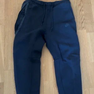 Nike tech byxor - Säljer ett par svarta mjukisbyxor från Nike med dragsko i midjan och sidofickor. Perfekta för en avslappnad stil eller träning. Byxorna har en enkel och stilren design med en liten Nike-logga på benet.