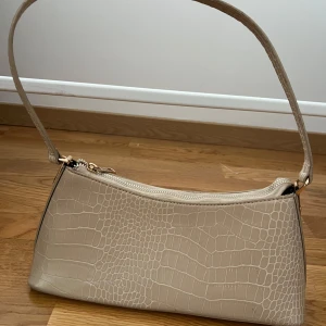 Beige axelväska med krokodilmönster - Snygg beige axelväska med krokodilmönster och dragkedja. Perfekt för att ge en stilren touch till din outfit. Väskan har en elegant design med ett bekvämt handtag.