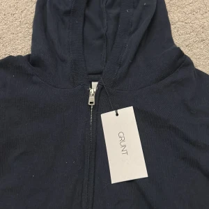 Mörkblå huvtröja från Grunt - Säljer en stilren mörkblå huvtröja från Grunt. Tröjan har en dragkedja framtill och en praktisk huva. Perfekt för en avslappnad look. Och helt ny tröja som aldrig har används 