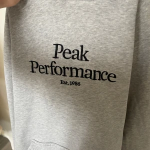 Grå hoodie från Peak Performance - Säljer en stilren grå hoodie från Peak Performance med broderad logga på bröstet. Tröjan har en klassisk känguruficka och ribbade muddar vid ärmsluten. Perfekt för en avslappnad stil.