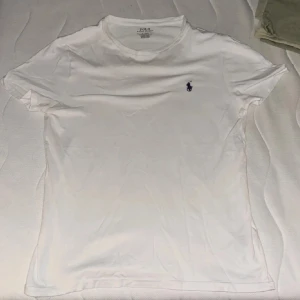 Vit t-shirt från Polo Ralph Lauren - Säljer en stilren vit t-shirt från Polo Ralph Lauren med den klassiska loggan broderad i blått på bröstet. T-shirten har en slim passform och är tillverkad i mjukt bomullsmaterial. Perfekt för en avslappnad och snygg look.