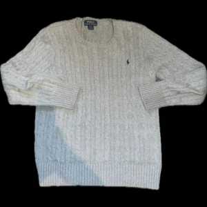 Grå stickad tröja från Polo Ralph Lauren - Säljer en stilren grå stickad tröja från Polo Ralph Lauren med kabelstickat mönster. Tröjan har långa ärmar och en rund halsringning. Perfekt för kyligare dagar.