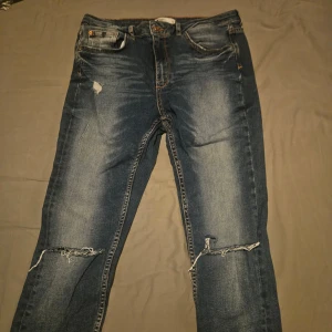 Blå jeans med slitningar - Säljer ett par blå jeans med coola slitningar på knäna och framsidan. De har en klassisk femficksdesign och knappgylf. Perfekta för en avslappnad stil.